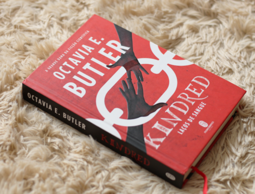 O livro tem uma capa dura vermelha com o nome da autora "Octavia E. Butler" e o título "Kindred" escritos em branco e também uma ilustração de duas mãos se tocando na ponta dos dedos e uma corrente branca ao fundo. O livro está sobre um tapete peludo bege.